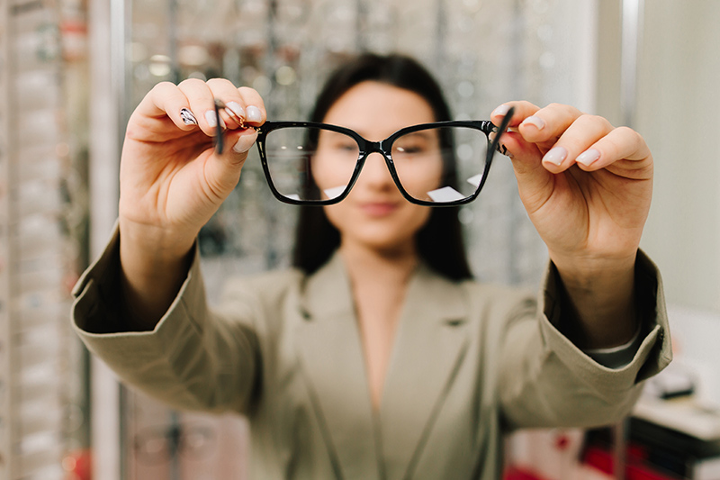 ผู้หญิงกำลังถือแว่นสายตาเลนส์โปรเกรสซีฟไว้ด้านหน้า เพื่อให้โฟกัสที่เลนส์ โดยมีฉากหลังเป็นร้านแว่นตา