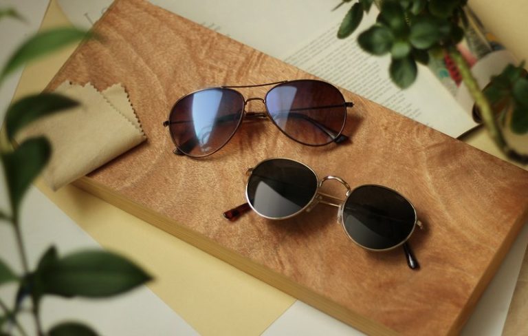 แว่นกันแดดทรงกลมและทรงนักบิน วางบนแผ่นไม้ พร้อมผ้าเช็ดเลนส์ บรรยากาศมินิมอล