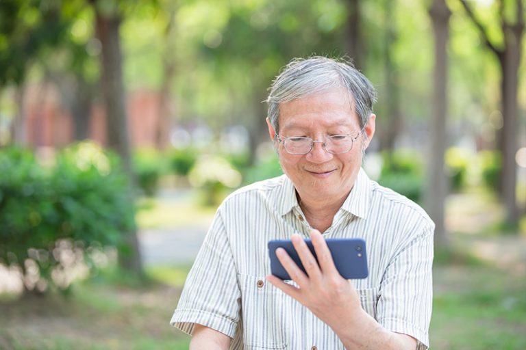 ผู้สูงอายุสวมแว่นกำลังยิ้มขณะใช้สมาร์ทโฟนในสวน
